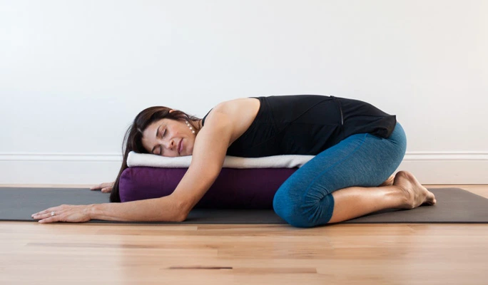 Mujer practicando Balasana o postura del niño en yoga con apoyo de bolster, variante restaurativa para mayor relajación.
