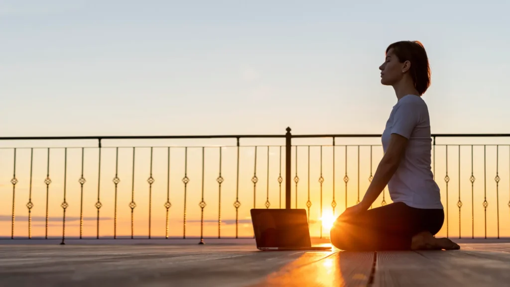 Mujer practicando mindfulness y meditación al amanecer frente a su computadora portátil en un espacio al aire libre.
