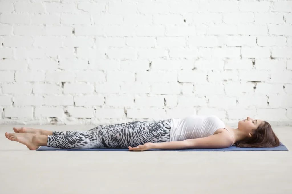 Mujer practicando la postura del cadáver o Savasana en yoga, tumbada boca arriba sobre esterilla azul con brazos extendidos y cuerpo completamente relajado.