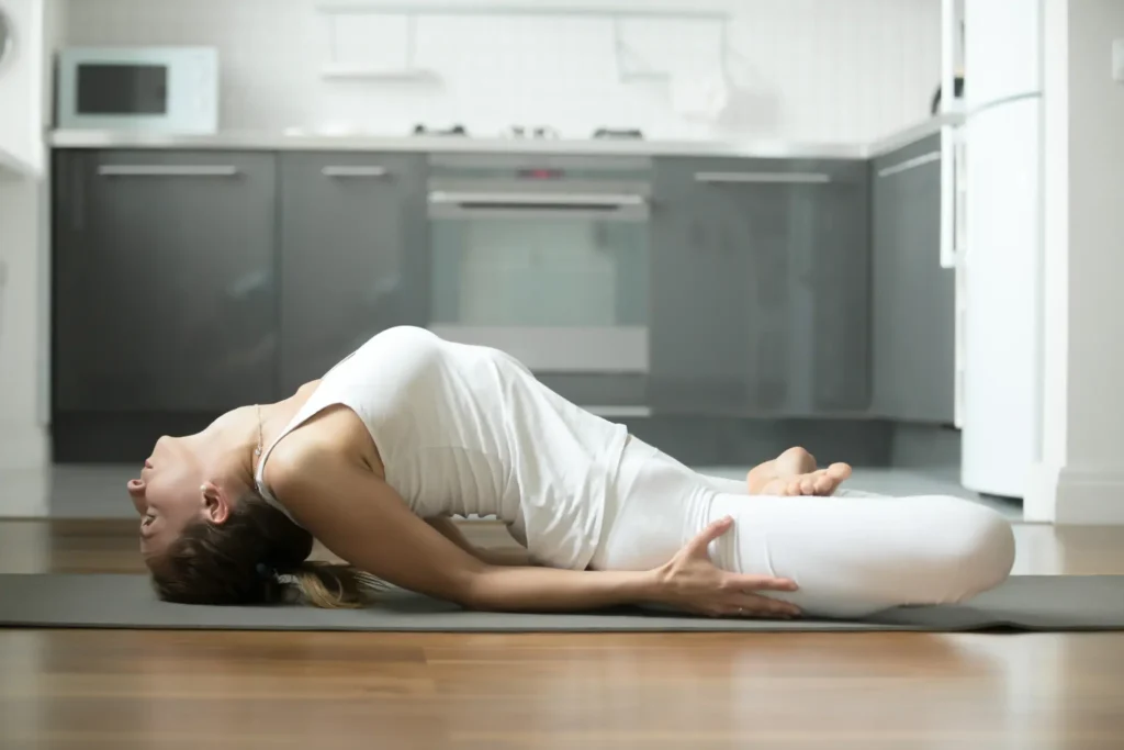 Mujer practicando la postura de la diosa reclinada Supta Baddha Konasana en casa sobre una esterilla de yoga.