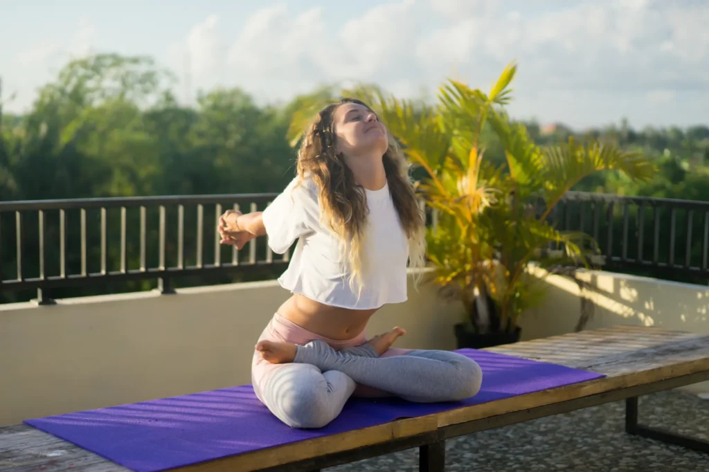 Mujer joven practicando yoga en posición de loto sobre una esterilla morada, estirando los brazos hacia atrás en una terraza al aire libre.