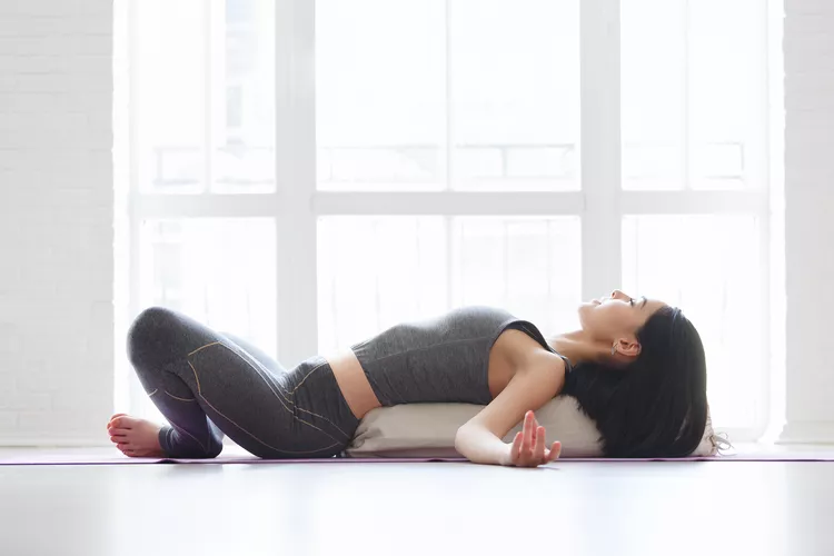 Mujer realizando Supta Baddha Konasana con apoyo de cojín, postura de yoga restaurativa para relajar el cuerpo y la mente
