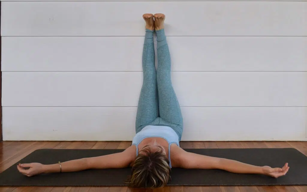 Mujer practicando Viparita Karani o postura de piernas en la pared, con brazos relajados en el suelo sobre un mat de yoga.